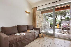 A brown sofa with a magazine, a straw hat, and a woven basket in a room with a stone wall and a glass door leading to an outdoor area with a table and chairs. - Maisonette in Gordes with Provençal Charm (GORDES)