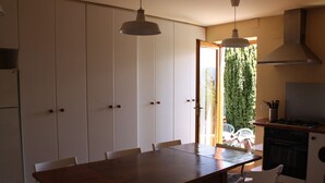 Un comedor con mesa de madera y sillas blancas, una cocina con estufa y lavavajillas, y vistas al exterior con vegetación.