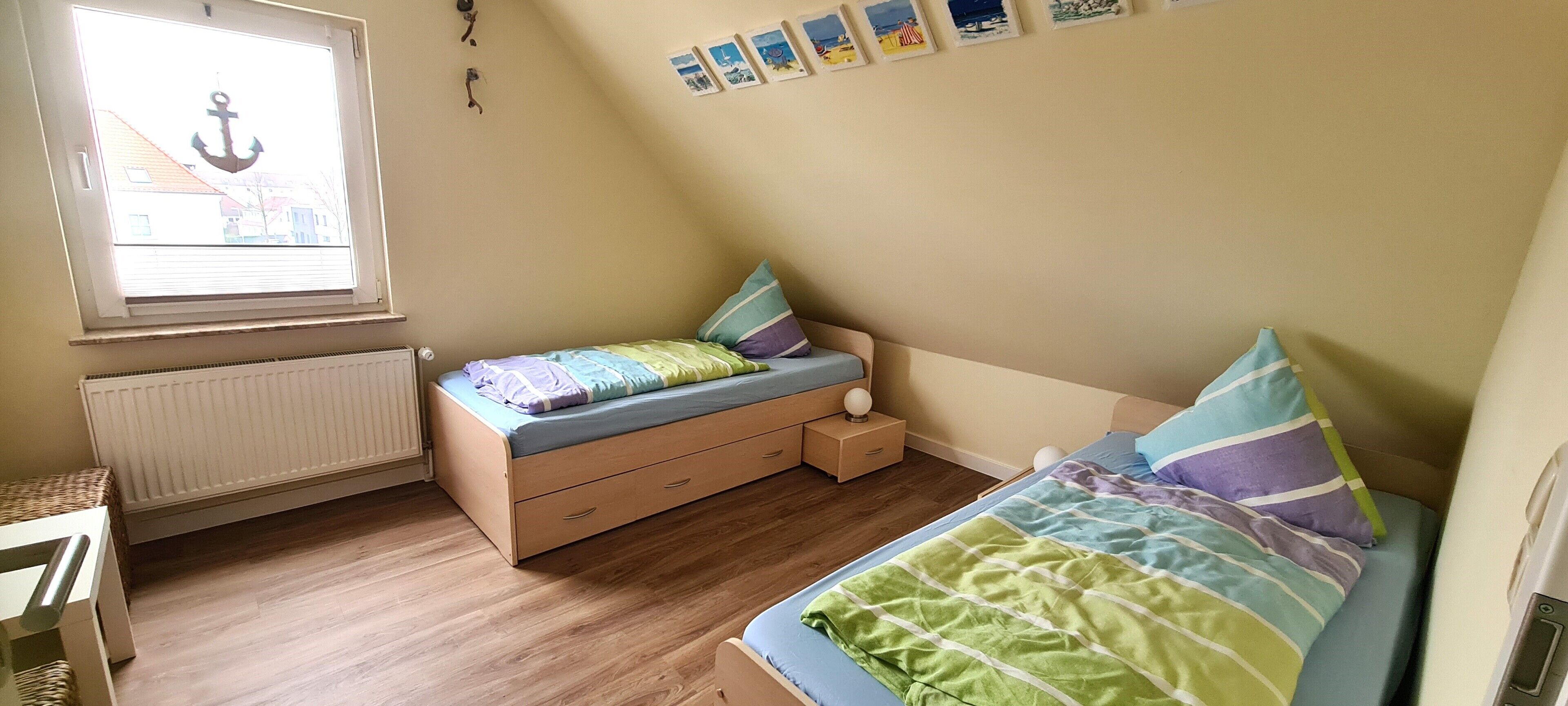 Ein Schlafzimmer mit zwei Betten, einem Fenster mit Anker-Dekoration, einem Heizkörper und einem kleinen Nachttisch mit Lampe.