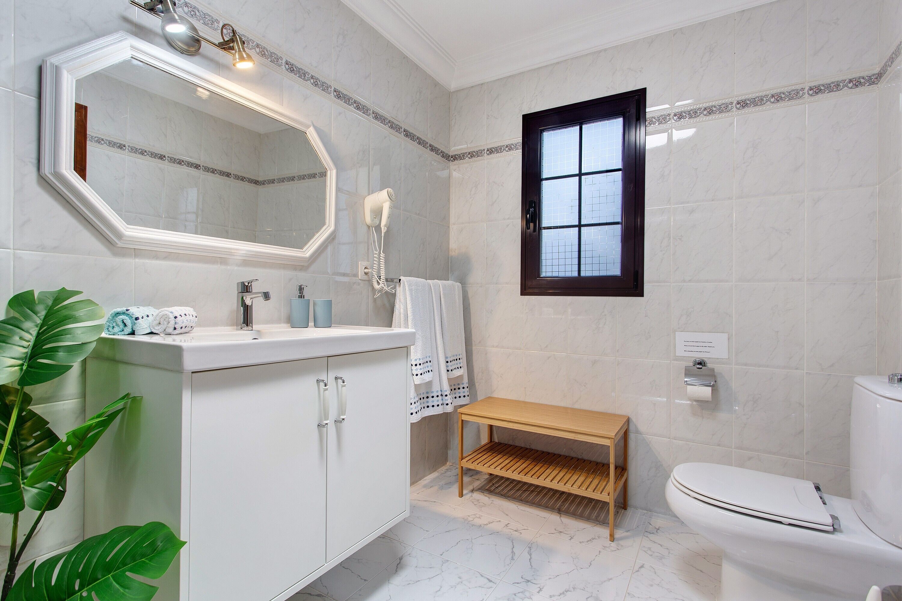 Un baño con tocador blanco, espejo, perchero, banco de madera, inodoro y ventana.
