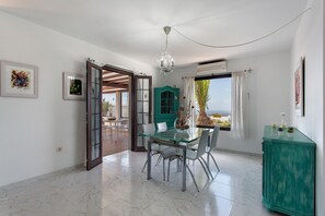 A dining area with a glass table, chairs, a chandelier, a teal cabinet, and a view through a window.