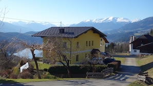 A yellow building with a dark roof, surrounded by a rural landscape with mountains in the background.