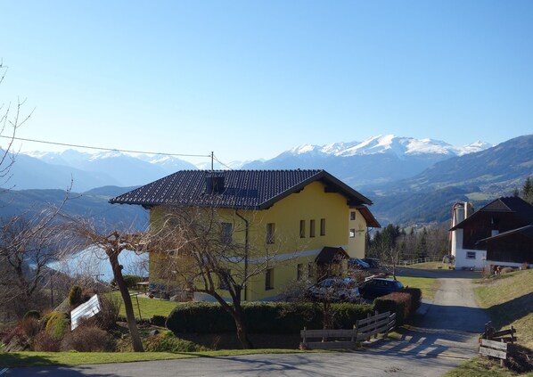 A yellow building with a dark roof, surrounded by a rural landscape with mountains in the background.