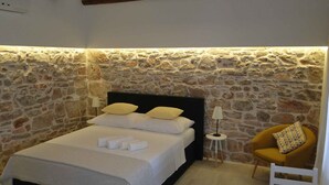 A bedroom with a stone wall, a bed with white linens, a small bedside table with a lamp, and a yellow armchair. - Studio with terrace and sea view (Primošten)