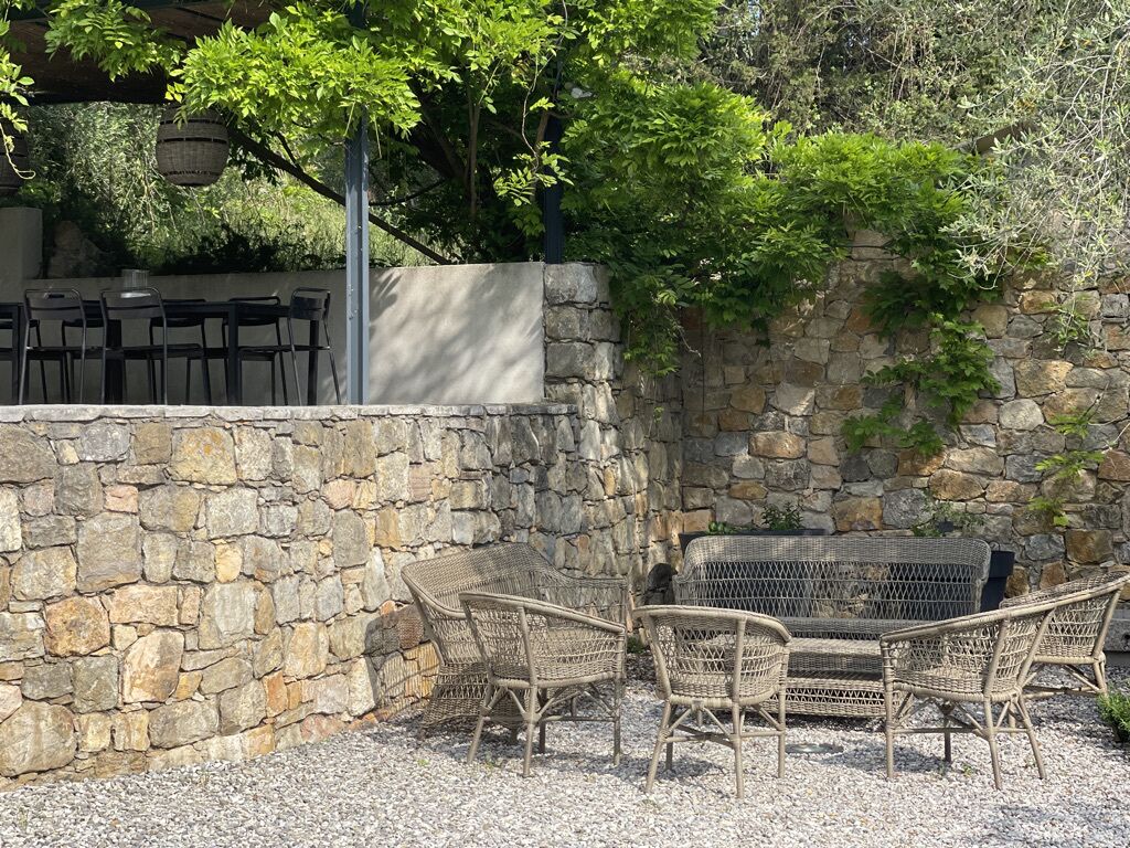 Un área de estar al aire libre con mobiliario de mimbre, una pared de piedra y una pérgola con follaje.
