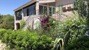 Ein zweistöckiges Haus mit Balkon, umgeben von üppigem Grün und blühenden Pflanzen.