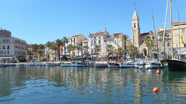 Ein Yachthafen mit vertäuten Booten, ein markanter Kirchturm und eine Reihe von Gebäuden mit Balkonen.