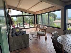 A sunlit patio with a wicker sofa, wooden chairs, a round table, and large windows offering a view of greenery and buildings.