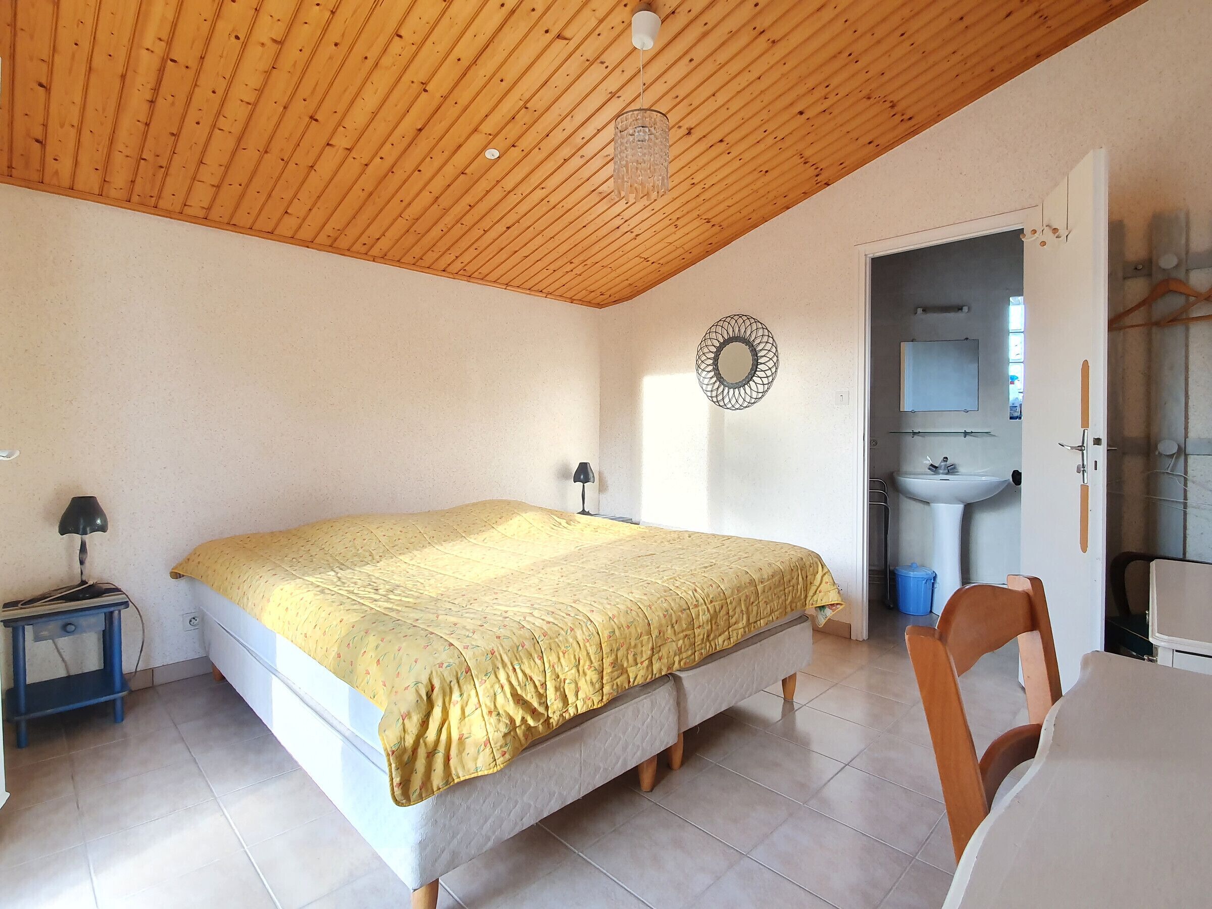 A bedroom with a wooden ceiling, a bed with a yellow bedspread, a bedside table with a lamp, and a bathroom visible through an open door.