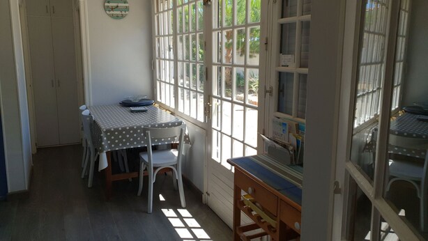 A room with a dining table, chairs, a clock, and a window with a view of greenery. - Charming renovated house, near beach and shops (La Faute-sur-Mer)