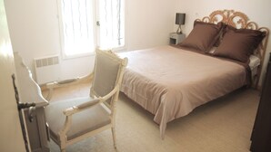 Quarto com cama, uma cadeira, mesa de cabeceira e janela com grades.