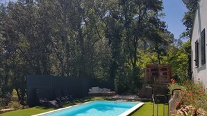 Uma piscina retangular com borda branca, cercada por grama sintética verde e um deck de madeira com um banco e uma mesa pequena.