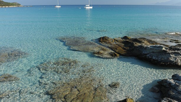 Mare turchese trasparente con rocce in primo piano e velieri all'orizzonte.