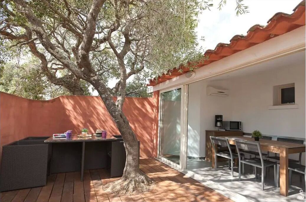 Eine Terrasse mit Esstisch und Stühlen, ein kleiner Tisch mit Gegenständen sowie ein großer Baum.