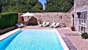 Una piscina rectangular con agua azul cristalina, rodeada por una terraza de madera. Se observan varias sillas de descanso y una mesa con sombrilla. Al fondo, se ve una pared de piedra y un seto.