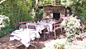 Un área de comedor al aire libre con un horno de piedra, rodeada de vegetación y una mesa preparada para una comida.