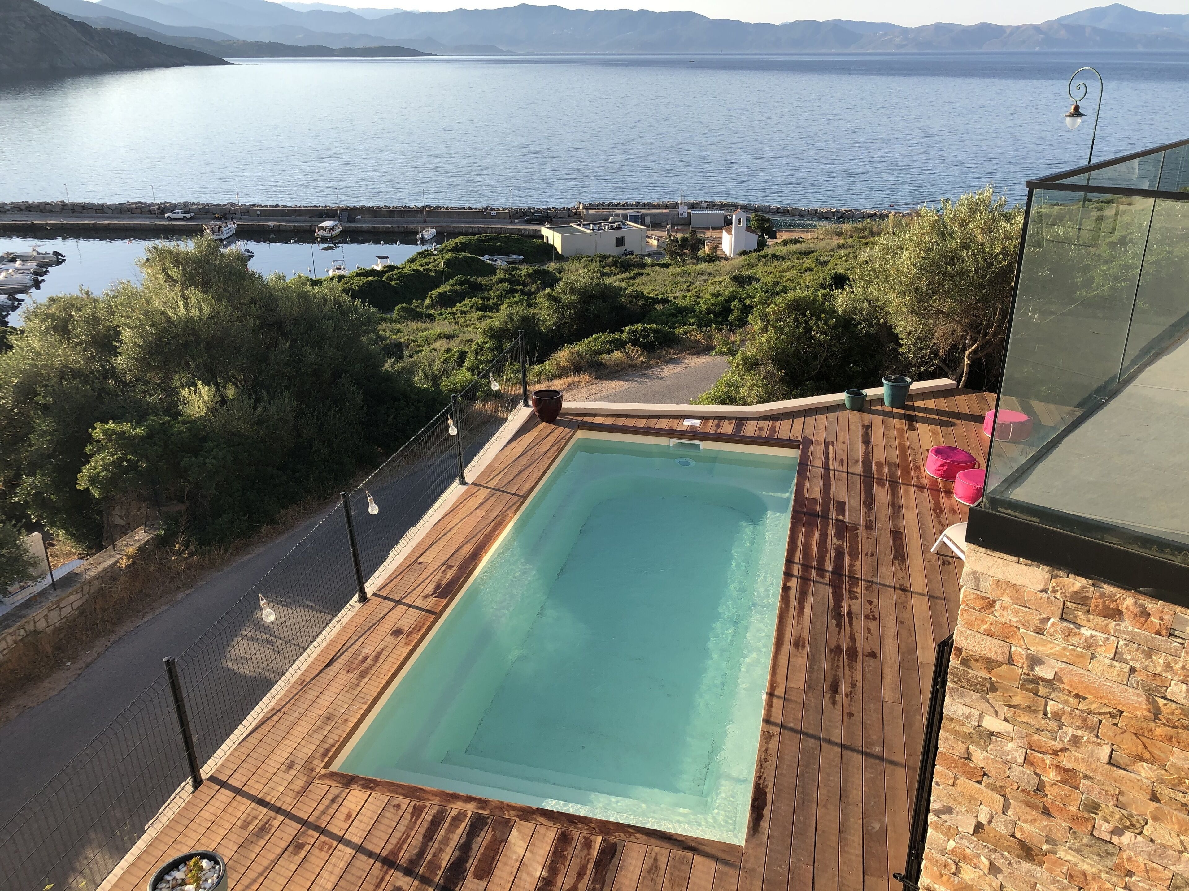A swimming pool with a wooden deck, surrounded by greenery and overlooking a body of water with boats.