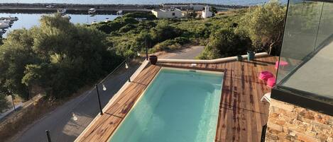 A swimming pool with a wooden deck, surrounded by greenery and overlooking a body of water with boats.