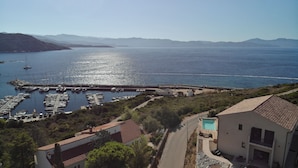 Un port de plaisance situé sur la côte, avec des bateaux amarrés, une piscine et une route menant à un bâtiment doté d’un toit en tuiles.