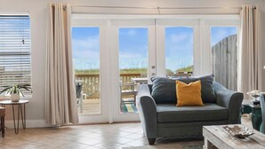 Sala de estar com sofá cinzento, almofada amarela e vista para a praia através de grandes portas de vidro.