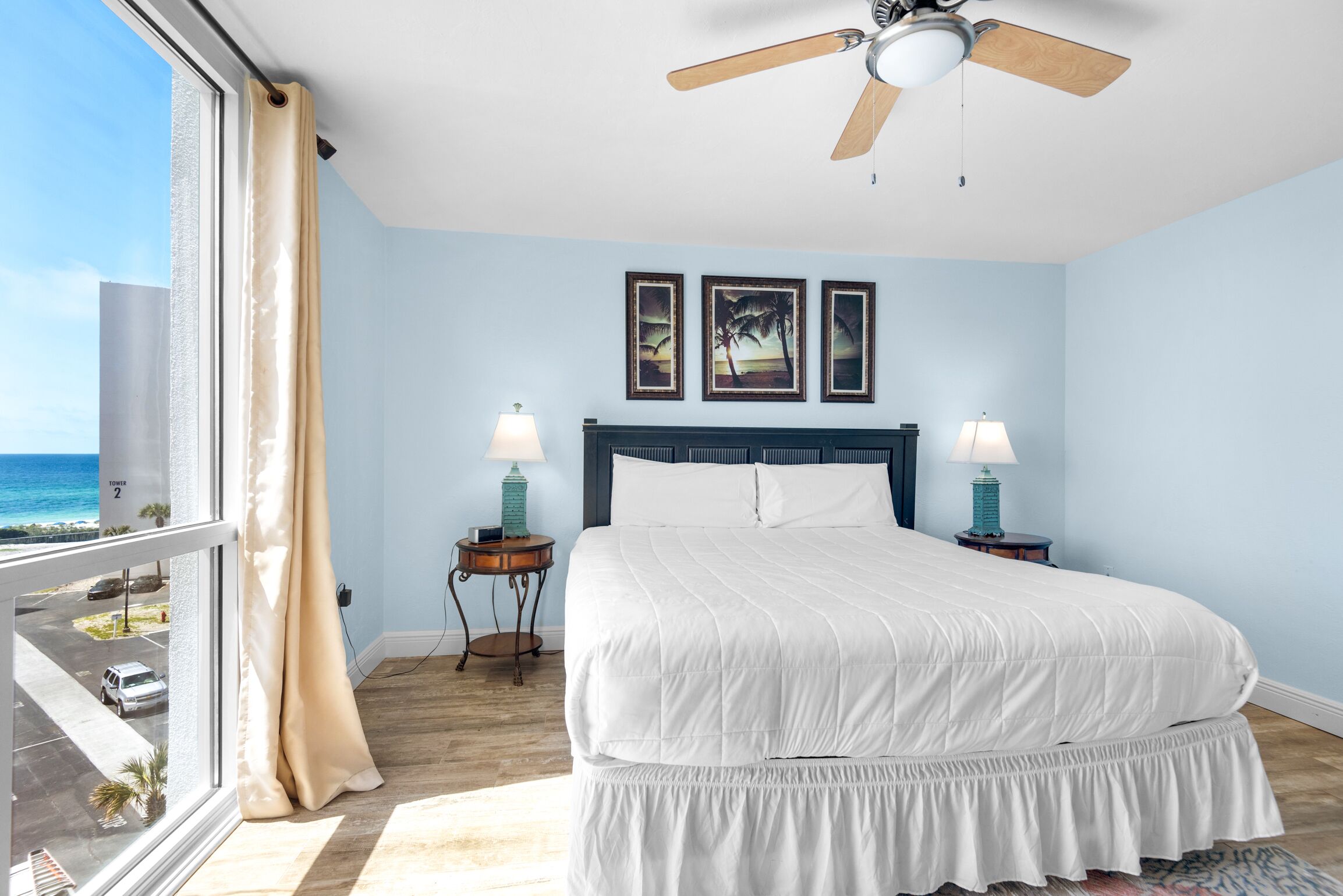 A bedroom with a large bed, two bedside lamps, a ceiling fan, and a view of the ocean.