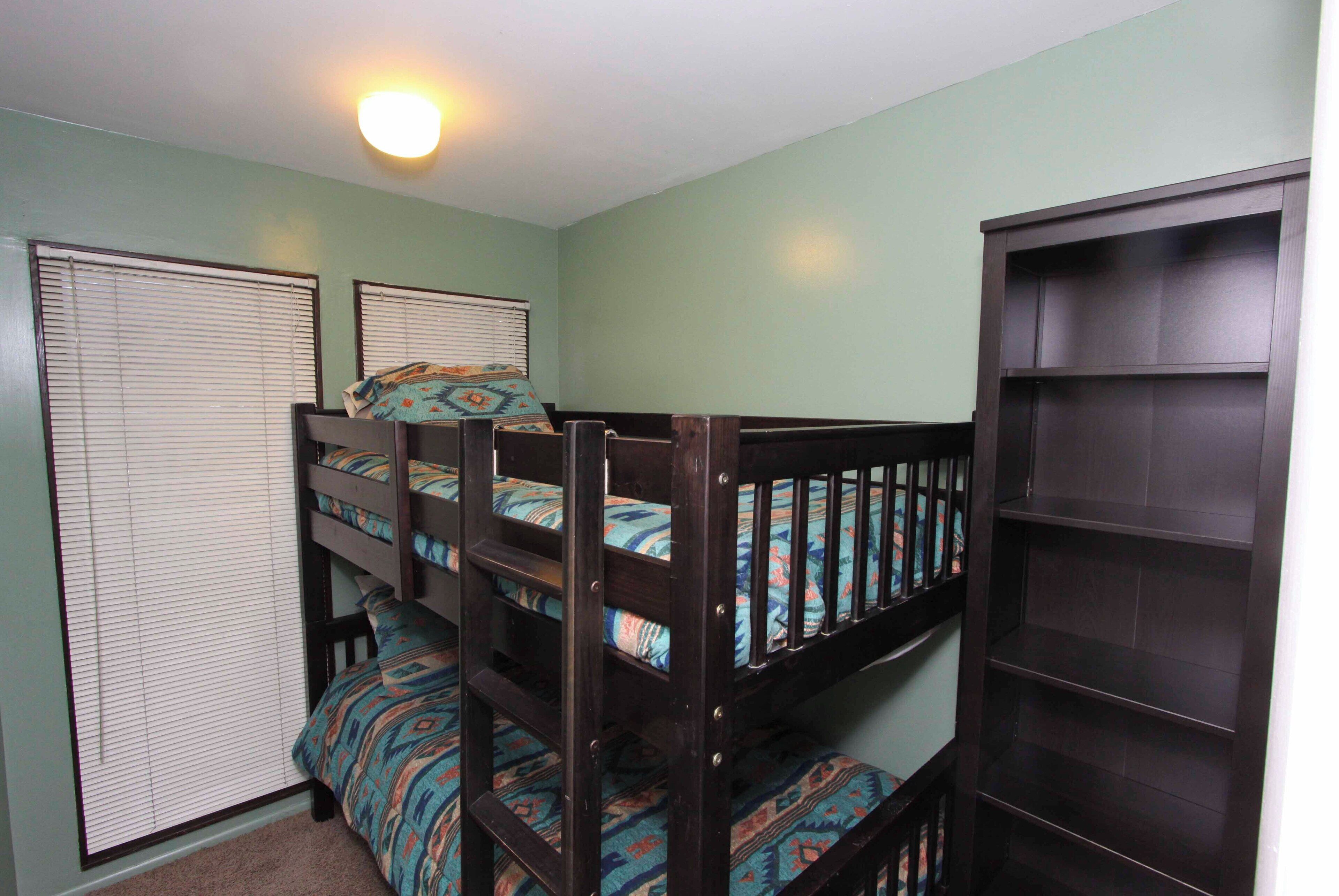 A bunk bed room with a dark wooden bunk bed, a small bookshelf, and a window with blinds.