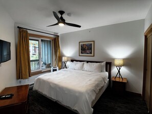 Habitación de hotel con cama, ventilador de techo, ventana con cortinas, televisor montado en la pared, lámparas de noche y un cuadro.