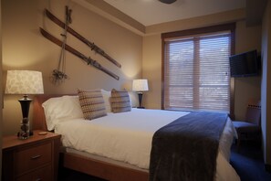 Quarto de hotel com cama, duas mesas de cabeceira com abajures, televisão na parede e janela com persianas.