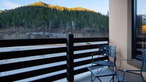 Een balkon met houten hekwerk, twee metalen stoelen en uitzicht op een bebost glooiend landschap.