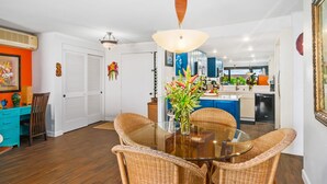 A dining area with wicker chairs and a glass table, a kitchen in the background, and a white door with a decorative item. - Breathtaking Panoramic Bali Hai with Unobstructed Bali Hai Ocean View (Princeville)