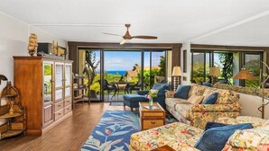 A living room with a sofa, a wooden cabinet, and a ceiling fan. There is a view of the outdoors through large windows. - Breathtaking Panoramic Bali Hai with Unobstructed Bali Hai Ocean View (Princeville)