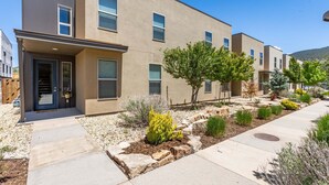 A modern multi-story residential building with a flat roof, large windows, and a landscaped front yard with various plants and a pathway.