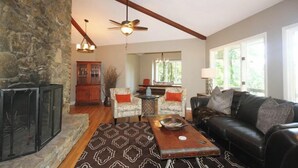 A spacious living room with a stone fireplace, a ceiling fan, and a large window providing natural light.