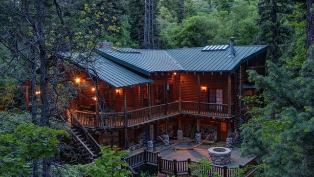 Una cabaña de troncos de dos pisos con balcón, rodeada de un bosque frondoso.