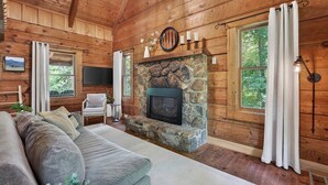 A log cabin interior with a stone fireplace, wooden walls, a sofa, and a television. - Falling Leaf Cabin Rustic Charm With Modern Decor! High Speed Internet!  (Rileyville)
