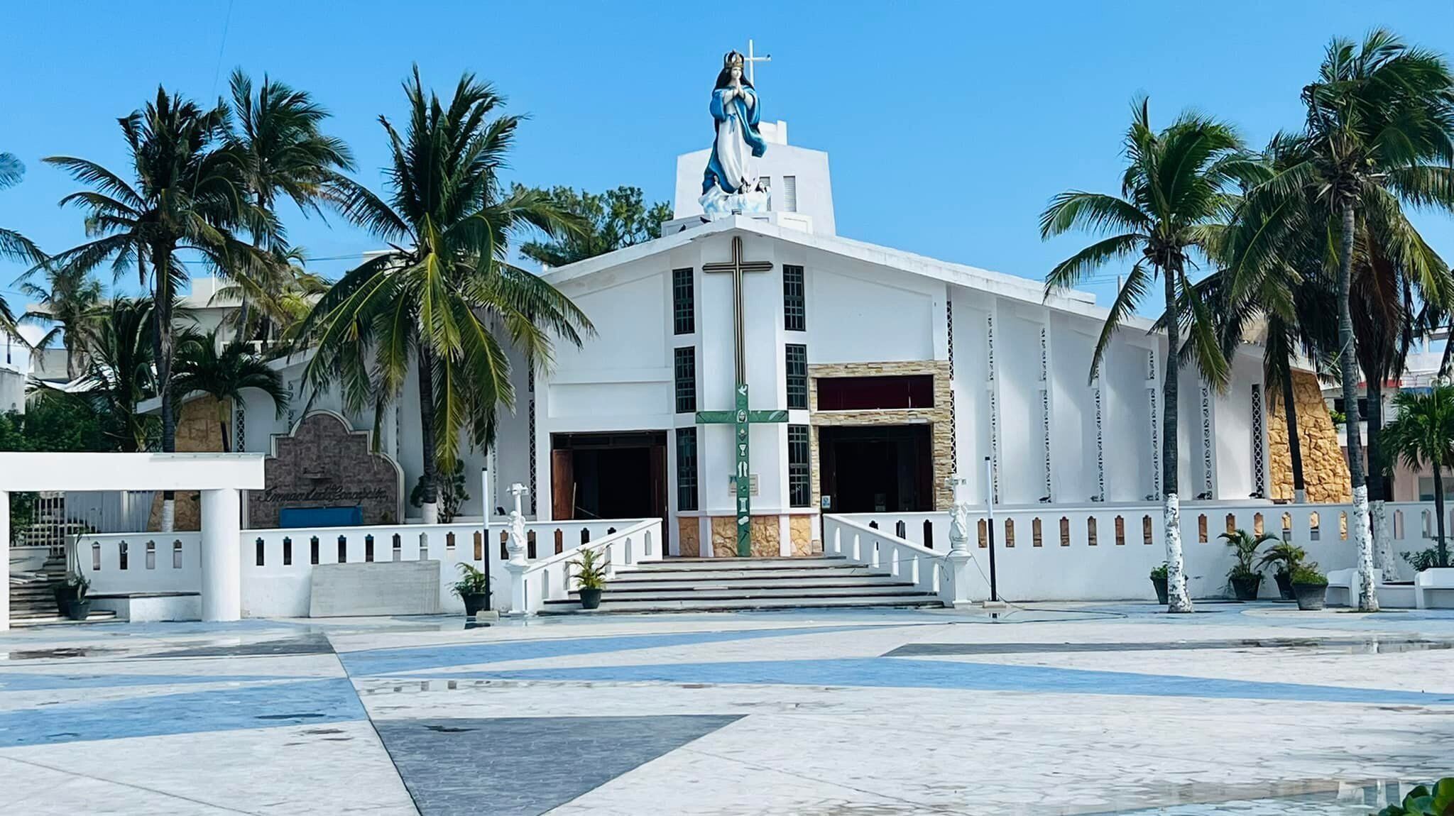 Una iglesia blanca con una estatua en la parte superior, palmeras y una cruz en el edificio.