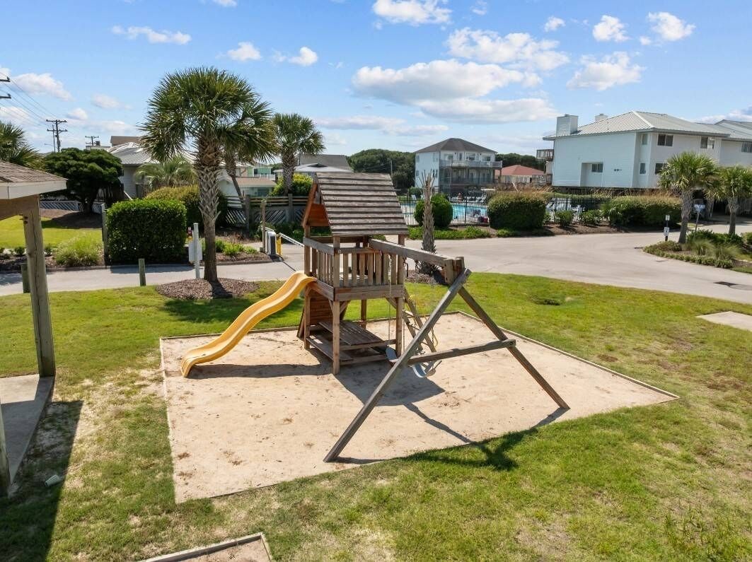 Un terrain de jeux avec un toboggan et des balançoires, situé dans un quartier résidentiel, avec des maisons et une piscine en arrière-plan.