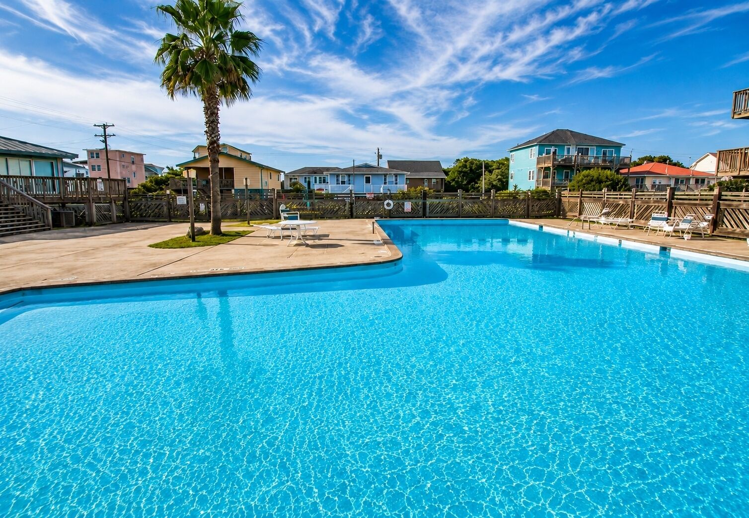 A swimming pool with clear blue water, surrounded by a wooden fence, and houses in the background.