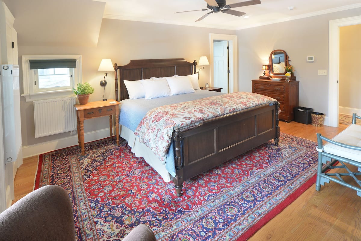 A bedroom with a large bed, a wooden dresser, a nightstand, a radiator, a window with a view, and a ceiling fan.
