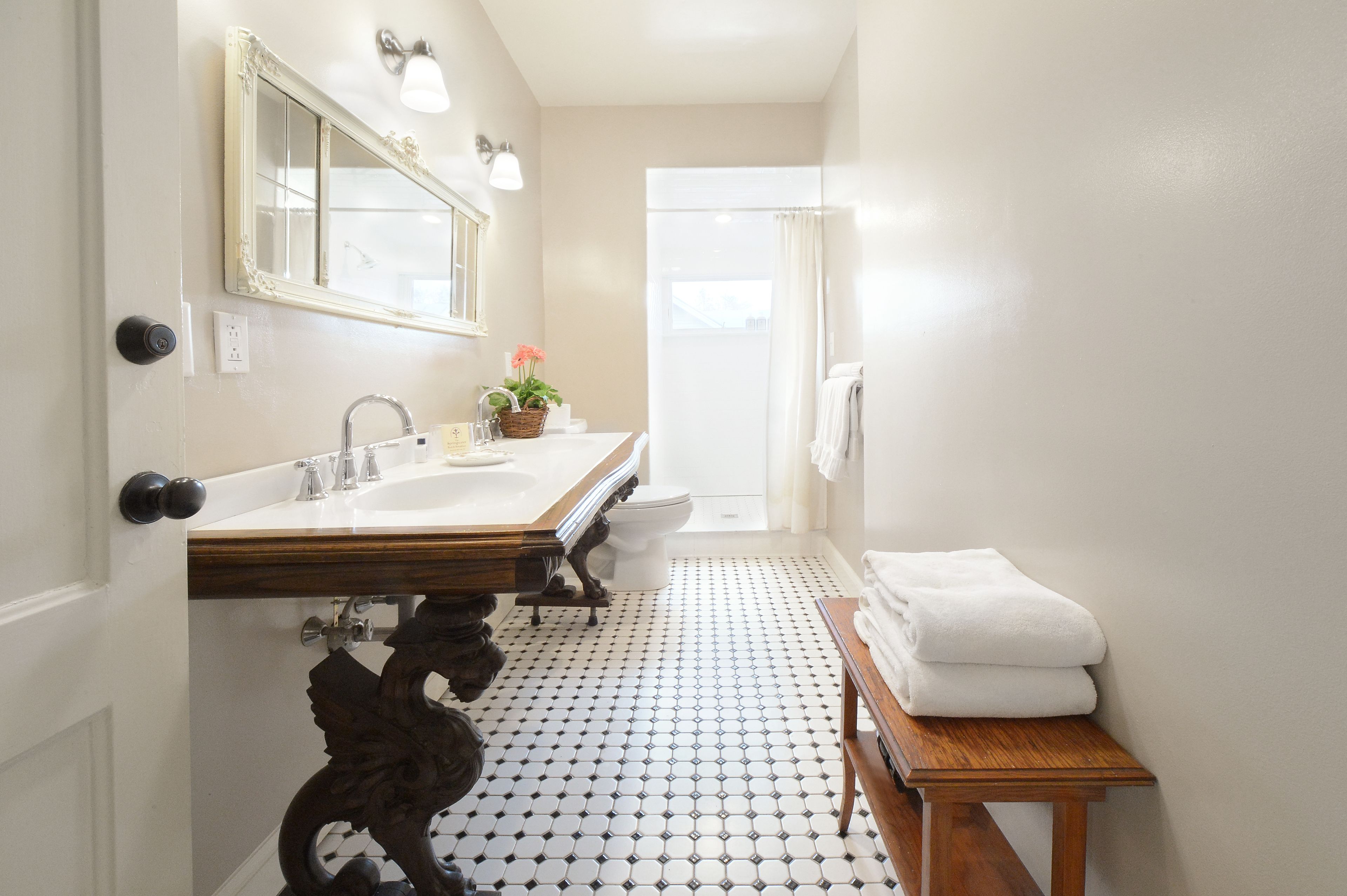 A bathroom with a white sink, mirror, and toilet. There is a wooden bench with towels and a potted plant on the sink.