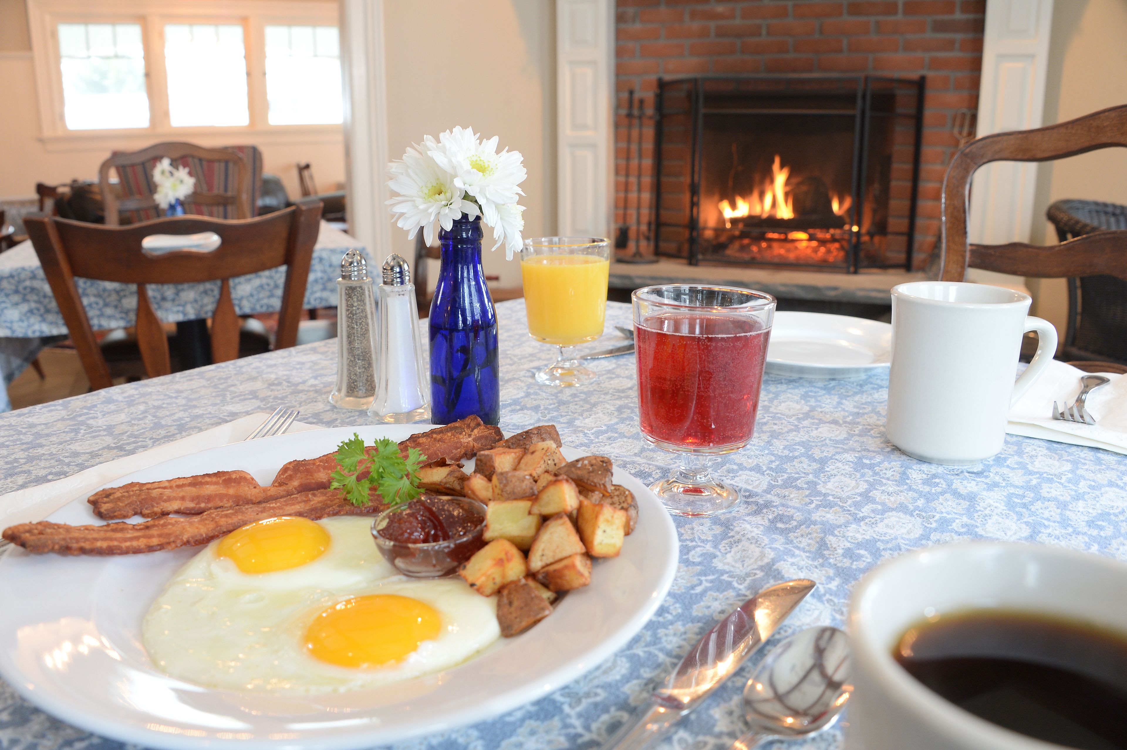 Une table dressée pour le petit-déjeuner avec une assiette d’œufs, de bacon et de pommes de terre, un verre de jus d’orange, un verre d’une boisson rouge et une tasse de café, le tout posé sur une table recouverte d’une nappe bleue à motifs.