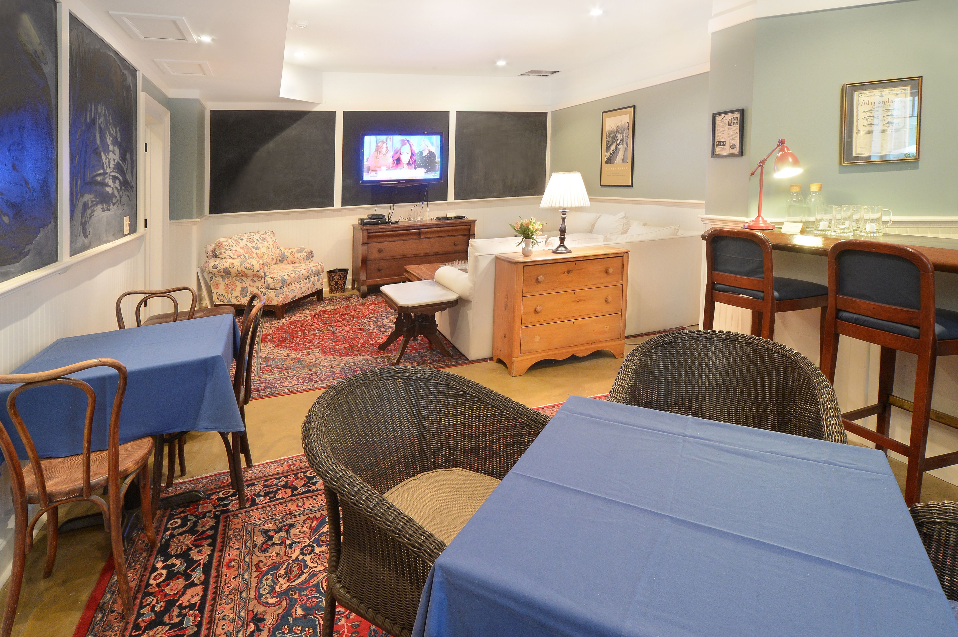 A room with a television, a desk with a lamp, wicker chairs, a sofa, and a dining table with blue tablecloths.