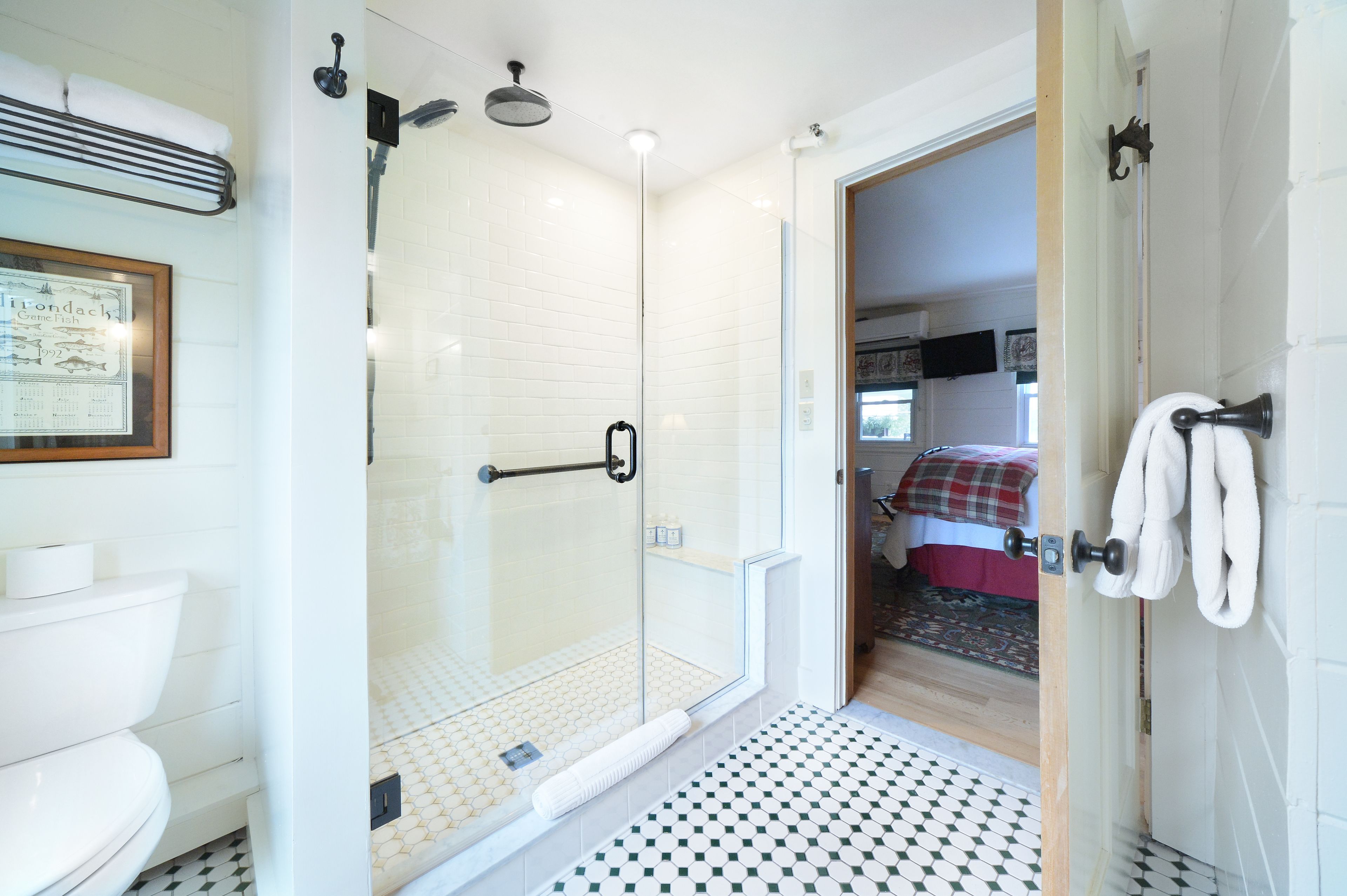 Un baño moderno con cabina de ducha de vidrio, inodoro y una habitación visible con cama y manta a cuadros rojos y blancos.