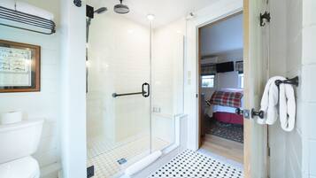 Un baño moderno con cabina de ducha de vidrio, inodoro y una habitación visible con cama y manta a cuadros rojos y blancos.