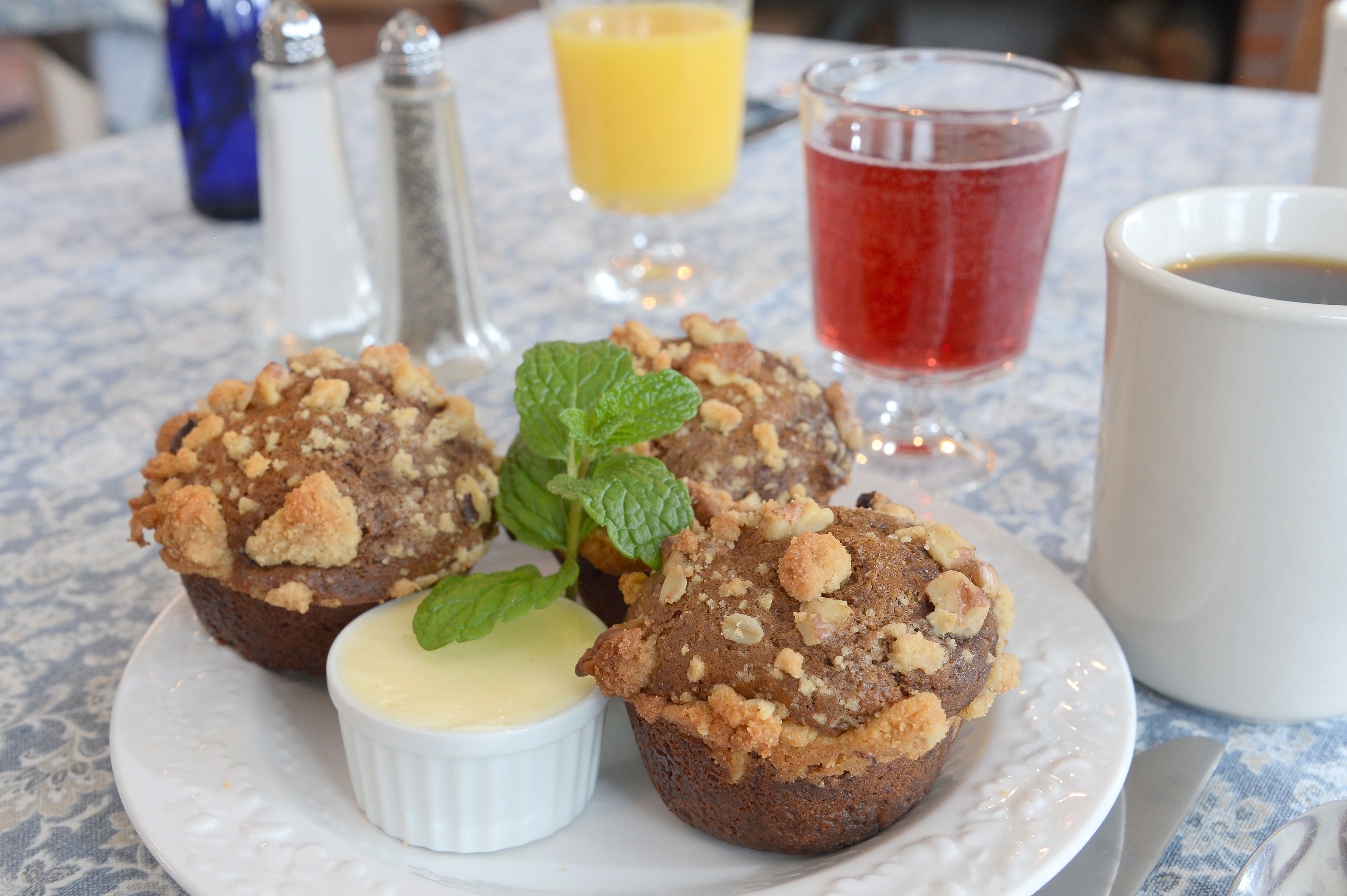 A plate with muffins and a small bowl of cream, a glass of red drink, a glass of orange juice, and a cup of coffee on a table.