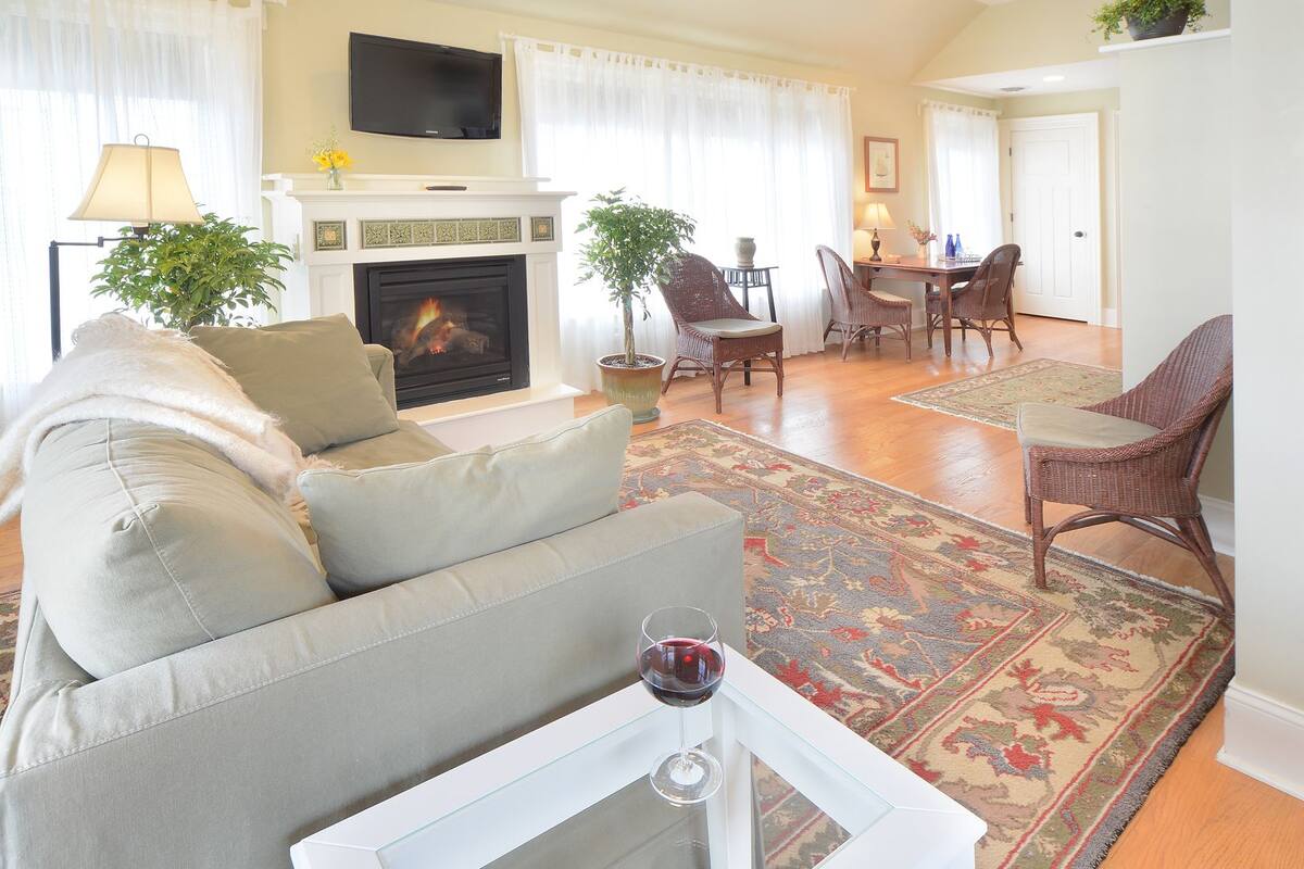 A living room with a fireplace, a television mounted above it, a dining area with chairs, and a glass table with a wine glass.