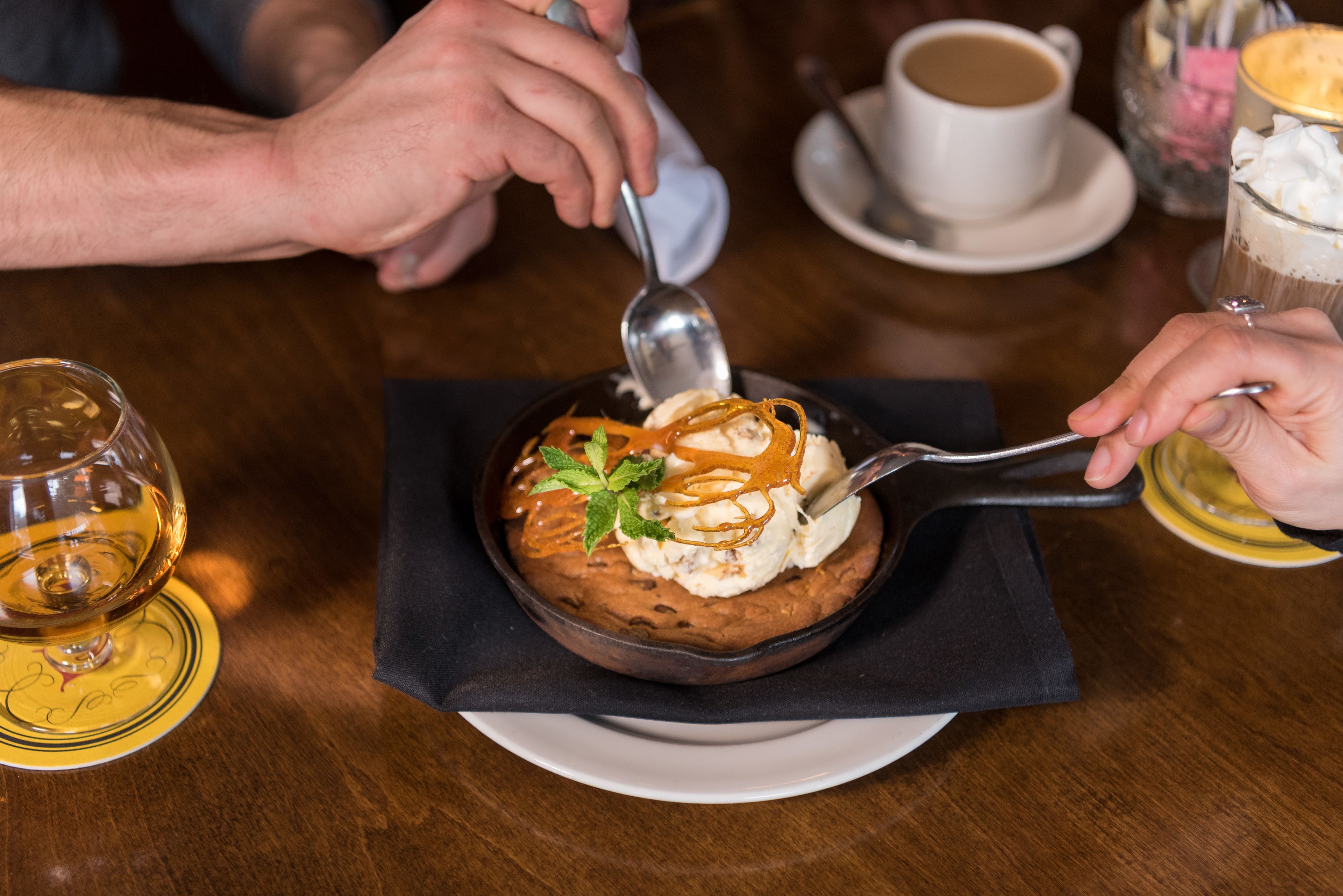 A dessert in a bowl with a spoon, a cup of coffee, and a glass of wine on a table.