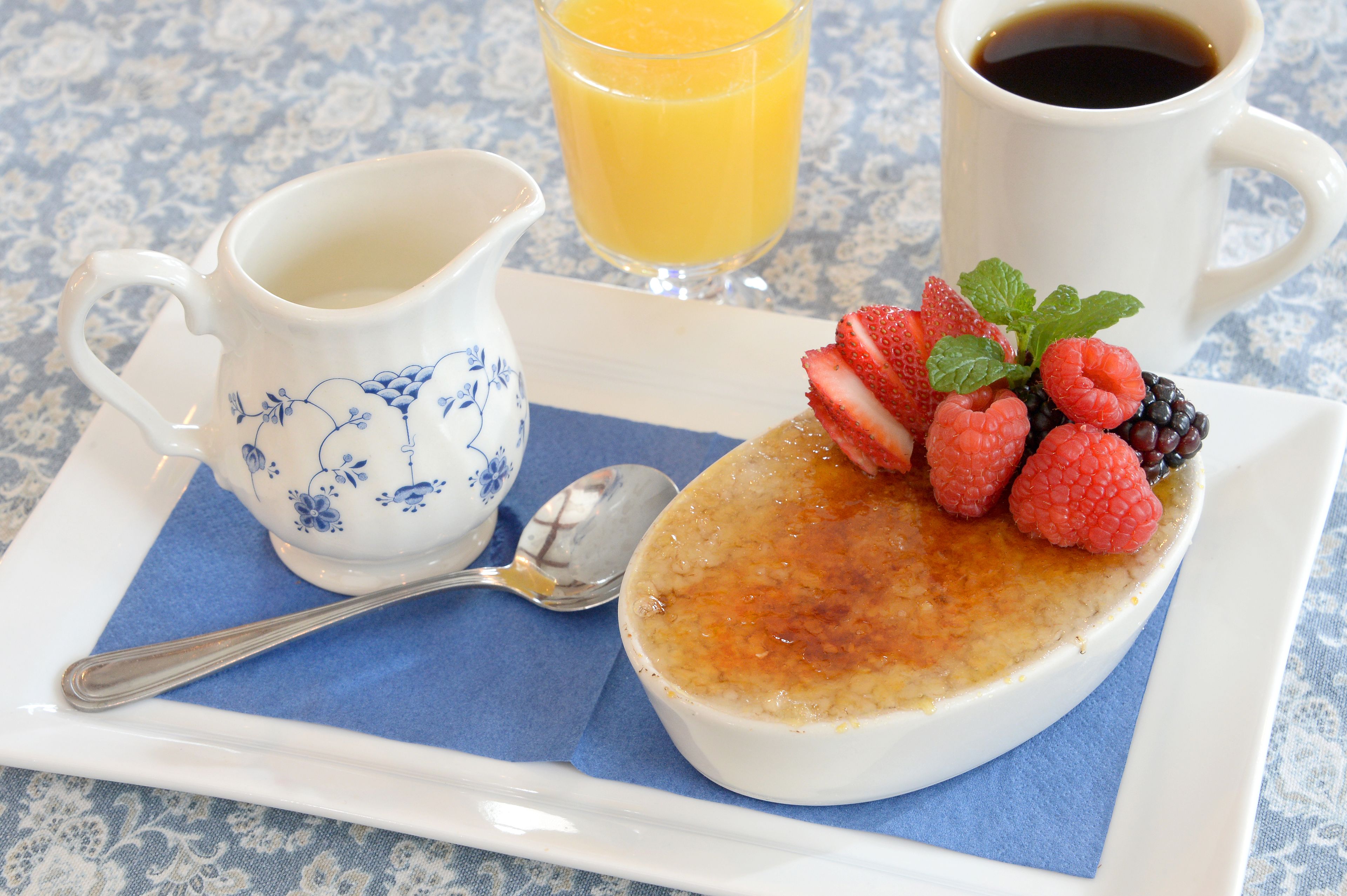 A dessert plate with crème brûlée, strawberries, raspberries, and blackberries, accompanied by a pitcher of cream, a glass of orange juice, and a cup of coffee.
