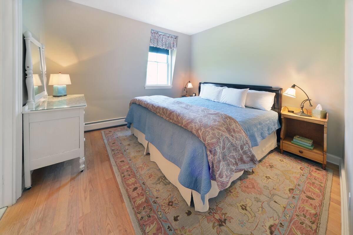 A bedroom with a bed, bedside tables, a nightstand, a mirror, a window, and a patterned rug.