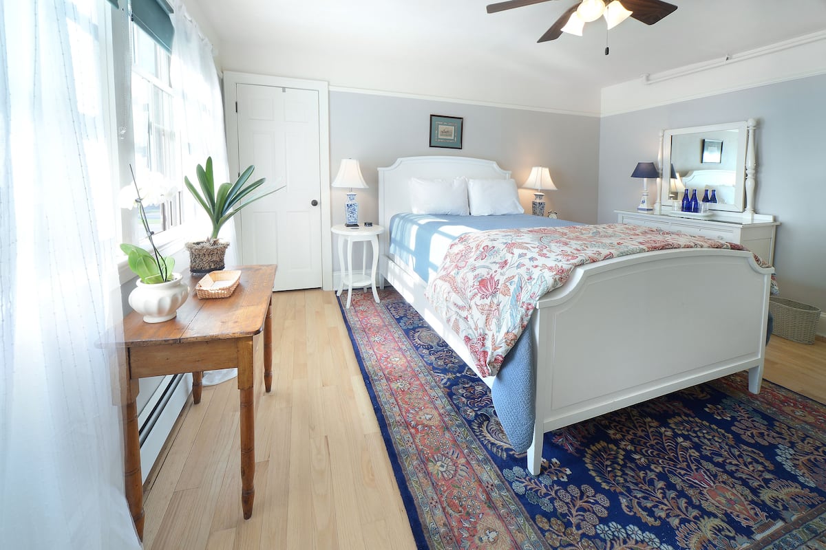 A bedroom with a bed, bedside tables, a desk, a potted plant, and a large window.